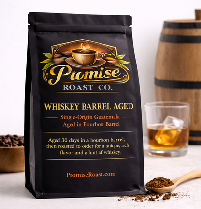 Black coffee bag from Promise Roast Co. with a glass of whiskey and wooden barrel in the background.